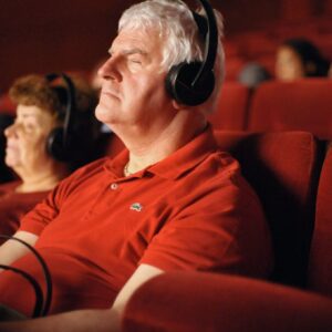 Older adults sitting in a dark theater wearing headphones, with eyes closed or relaxed. The man in the foreground, dressed in a red shirt, appears to be immersed in an audio-described experience. The setting is calm and inclusive.
