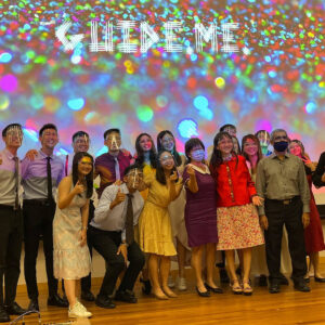A diverse group of people posing happily on a stage in front of a colorful, sparkling background with the words "Guide Me" displayed above. The individuals are dressed in semi-formal to formal attire, celebrating at what appears to be a festive or inclusive event.