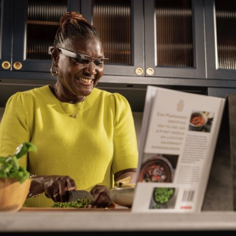 A smiling woman wearing smart glasses is chopping vegetables in a modern kitchen. She is following a recipe from an open cookbook on the counter.