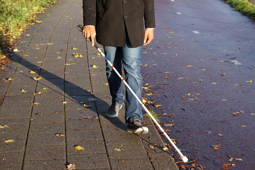 AdobeStock_231187109-web_835x555_opt A visually impaired man walks along a sidewalk using a long white cane for navigation. He is wearing jeans, a black coat, and dark shoes, and is walking near the edge of a wet pavement scattered with fallen leaves.